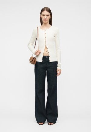 Kvinne iført en hvit, langermet cardigan med knapper, delvis åpen, svarte vide bukser, svarte sandaler og en liten brun skulderveske.