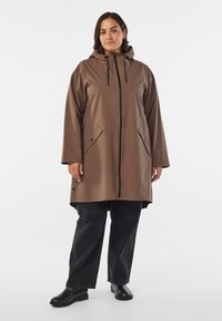 Bruine waterdichte jas met een capuchon en een ritssluiting aan de voorkant, voorzien van zijzakken en verstelbare trekkoorden. Gepaard met zwarte wijde broek en laarzen.