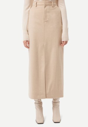 Frau trägt einen beigen hoch taillierten Midirock mit einer vorderen Naht, kombiniert mit einem langärmligen Oberteil und beigen Stiefeletten.