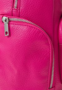 Sac à dos en cuir rose vif avec deux fermetures éclair argentées visibles sur les sections avant et supérieure courbées, montrant une texture et des coutures détaillées.