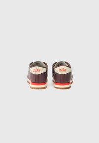 Nike-kengät tumman violetista nahasta, joissa on valkeat ja punaiset yksityiskohdat. Ominaisuuksina pyöristetty muoto, tasapohja ja näkyvät saumat. Logo kantapäässä.