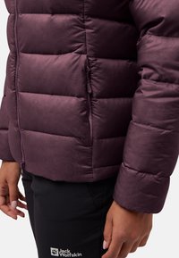 Primo piano di una giacca trapuntata viola con zip e tasca laterale, indossata con pantaloni neri Jack Wolfskin.