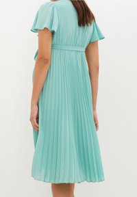 Světle tyrkysové skládací šaty s lehkými rukávy, pasem na šňůrku a splývavou tkaninou, dosahující těsně pod kolena, s vertikálními sklady.