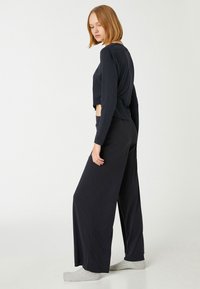 Mujer con pelo rubio corto que lleva una camiseta de manga larga negra y corta, pantalones negros anchos y calcetines grises, de pie de lado sobre un fondo blanco.