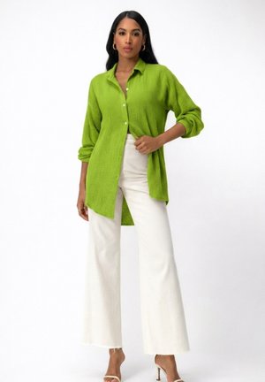 Mujer llevando una camisa de manga larga de textura verde brillante, pantalones blancos de pierna ancha y sandalias de tacón beige sobre un fondo blanco.