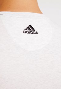 Camiseta blanca de algodón con cuello redondo, que presenta un logo negro de Adidas en la parte superior de la espalda. La tela tiene una textura sutil y suavidad.