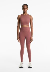 Woman standing, wearing mauve high-waisted leggings and matching cropped tank top, with white athletic shoes, on white background.