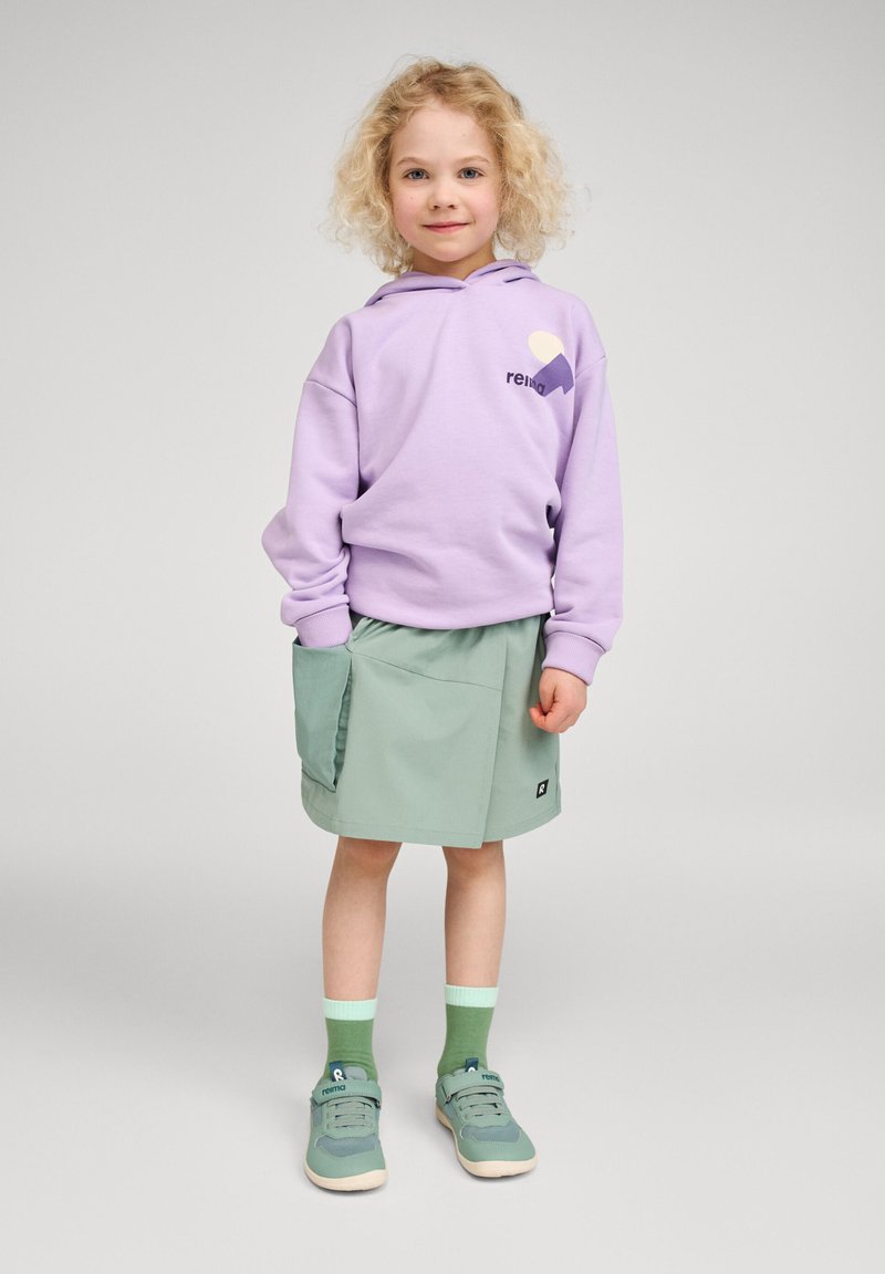 Παιδί με σγουρά ξανθά μαλλιά, όρθιο, φορώντας φούτερ με κουκούλα σε λιλά χρώμα, φούστα σε μέντα με τσέπη, πράσινες κάλτσες και τυρκουάζ αθλητικά παπούτσια σε απλό φόντο.