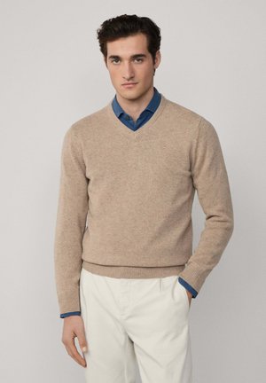 Beige V-hals trui van zacht breimateriaal, gedragen over een blauwe overhemd met kraag. De pasvorm is smal, met geribde boorden en zoom.
