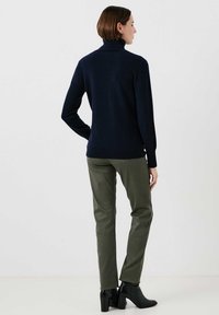 Jersey de cuello alto azul marino con mangas largas, combinado con pantalones verdes oliva y botas negras hasta el tobillo, mostrando una silueta estilizada.