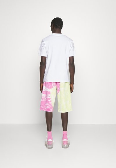 Fehér pamut póló, élénk rózsaszín és zöld batikolt rövidnadrág, rózsaszín zoknik és átlátszó tornacipők láthatók hátulról, semleges háttér előtt.