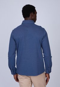 Chemise bleue à manches longues en tissu texturé avec un col clair et des poignets boutonnés, vue de dos sur un fond clair.