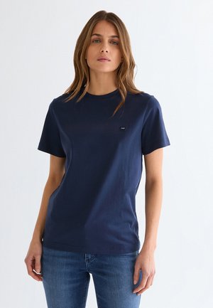 Vrouw met lichtbruin haar, gekleed in een effen donkerblauw T-shirt met korte mouwen en middelblauwe spijkerbroek, staand tegen een witte achtergrond.