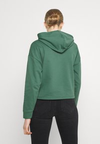 Žena s vlasy ve svázaném drdolu, oblečená v zelené mikině s kapucí a černých přiléhavých džínách, zobrazená zezadu na jednoduchém bílém pozadí.