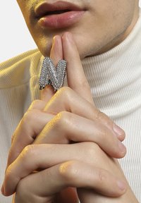 Sølv ring besat med rhinsten i form af bogstavet "N," båret på en finger, imod en lys baggrund med en ribbenstrikket turtleneck synlig.