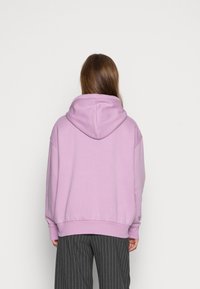 Sudadera con capucha de color lila y corte holgado, de tela texturizada y puños acanalados. Llevada sobre pantalones oscuros de rayas, mostrando la parte de atrás.