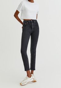 Jeans negros de tiro alto en denim con un ajuste slim, cinco bolsillos y detalles en botón de cobre, combinados con una camiseta corta blanca y zapatillas.