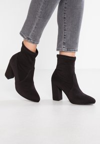 Botines negros de tacón alto hechos de suave ante. Presentan una punta puntiaguda y un tacón en bloque de altura media. El diseño incluye acentos cosidos.