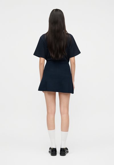 Γυναίκα με μακριά σκούρα μαλλιά που φοράει μπλε μπλουζάκι και φούστα, λευκές κάλτσες και μαύρα μοκασίνια, στέκεται γυρισμένη με την πλάτη σε λευκό φόντο.