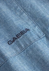 Lichtblauwe stof met een gestructureerde uitstraling. Bevat een geborduurd "GABBA" logo in marineblauw op de zak. Heldere stiksel details zichtbaar.