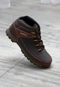 Bota de senderismo de piel marrón con suela de goma resistente, ojales dorados y logo de Timberland. Presenta un cuello acolchado y acentos texturizados.