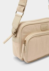 Beige színű steppelt keresztpántos táska, dombornyomott logóval, texturált pánttal és két cipzáras rekesszel a funkcionális tárolás érdekében.