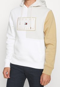 Uomo in felpa bianca con manica destra beige e logo Tommy Hilfiger sul petto, abbinata a pantaloni a quadri scuri.