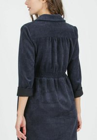 Marineblaues Cordkleid mit Kragen, gerafftem Rückenteil, elastischem Taillenbund und hochrollbaren Ärmeln. Weicher Griff mit vertikalen Rippungen.
