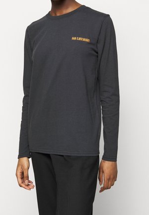 Osoba ubrana w dopasowaną, długorękawową ciemnoszarą koszulę z małym pomarańczowym napisem „HAN KJØBENHAVN” na piersi, w połączeniu z czarnymi spodniami.