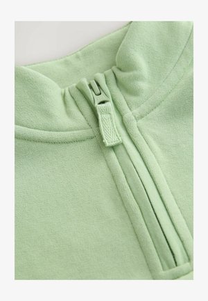 Svijetlozelena majica s visokim ovratnikom, koja ima zatvaranje na pola patentnog zatvarača i meku, teksturiranu tkaninu. Detalji šavova su vidljivi duž rubova.