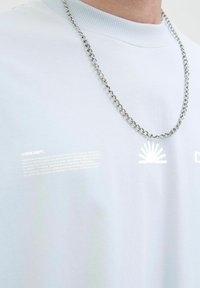 Himmelblaues Baumwoll-T-Shirt mit geripptem Halsbündchen; zeigt reflektierenden weißen grafischen Text und Sonnenmotiven auf der Vorderseite; silberne Halskette sichtbar.