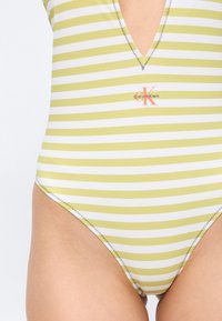 Ριγέ κίτρινο και λευκό μαγιό Calvin Klein με βαθύ V ντεκολτέ και ορατή ζιγκ-ζαγκ ραφή στις άκρες.