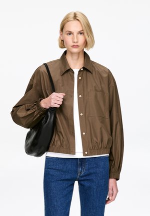 Kobieta o krótkich blond włosach nosi brązową zapinaną na guziki kurtkę, białą koszulę, niebieskie dżinsy i trzyma czarną torebkę na ramię na białym tle.