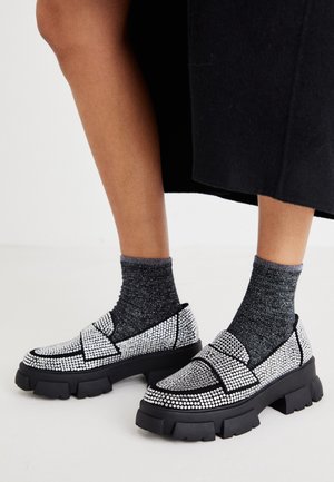 Sorte og sølv slip-on sko med rhinsten dekorationer, chunky såler og et struktureret sort accentdesign. Båret med mørke, glitrende sokker.