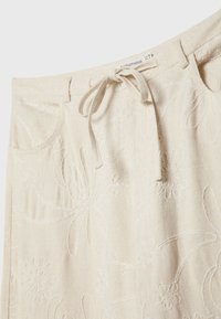 Jupe beige brodée avec des motifs floraux, lien de taille ajustable et deux poches latérales. Tissu texturé avec une finition douce.