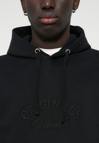 Hoodie noir avec un cordon de serrage, ornée de broderies "ORIGINALS" et "Studio", fabriquée dans un tissu doux et texturé.