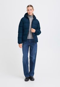 Mornarska puffasta jakna s kapuco, z značilnim prešitim dizajnom, zadrgo in rebrastimi manšetami, v kombinaciji s sivo turtleneck majico in modrimi denim hlačami.