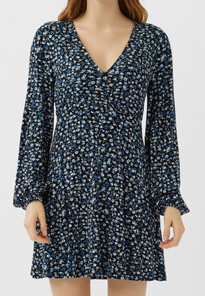 Vrouw die een zwarte jurk draagt met een kleine blauwe en witte bloemenprint, lange mouwen en een V-hals, staand tegen een effen achtergrond.