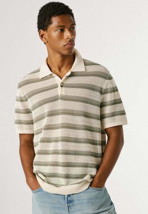 Jauns vīrietis, tērpies īsroku, apkaklētā trikotāžas krekliņā ar horizontālām zaļām un bēšām svītrām, ar rokām gaišu zilu džinsu kabatās, skatās labi.