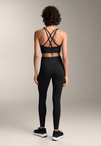 Fekete sportbra keresztezett pántokkal, magas derekú fekete leggingsekkel és fekete sportcipőkkel párosítva. Simább anyag, testhezálló dizájn.