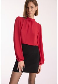 Czerwona bluzka z długim rękawem i marszczonym dekoltem. Połączona z czarną mini spódnicą. Tekstura wydaje się miękka; bluzka ma lekki połysk.