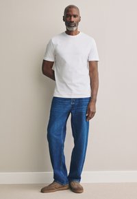 T-shirt bianca a maniche corte, jeans di denim blu, scarpe casual color cuoio. Il modello è in piedi con una mano dietro la schiena contro un muro neutro.