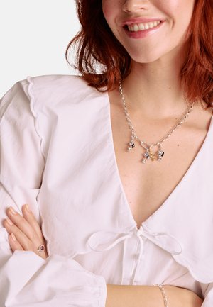 Silberne Kettenhalskette mit bunten Anhängern, die ein Blumen- und Blätterdesign zeigen. Das Modell trägt eine weiße, strukturierte Bluse mit einem gewellten Kragen.