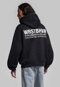 Felpa nera con una grande grafica bianca sulla schiena, dotata di spalle rotonde e una vestibilità ampia. Abbinata a jeans in denim azzurro chiaro.