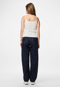 Donna in piedi a favore di spalle, che indossa un top bianco senza maniche, jeans a gamba larga blu scuro e sneakers bianche su uno sfondo uniforme.