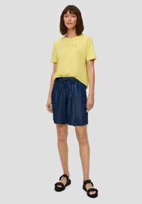T-shirt en coton jaune avec le texte "LOVELY", associé à un short en denim bleu foncé avec une taille élastique et des sandales noires à brides.
