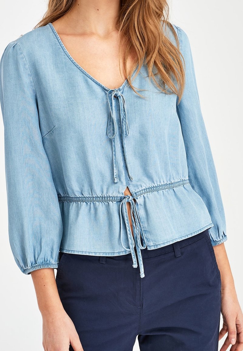 Hellbluse aus Denim, V-Ausschnitt mit Kordelzug, geraffter Taillensitz und Puffärmeln. Glatte Textur mit subtilen Nähdetails.