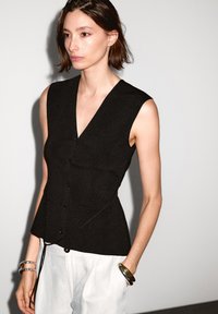 Gilet senza maniche nero con scollatura a V profonda, caratterizzato da bottoni lungo il davanti e cuciture strutturate. Indossato con pantaloni chiari e braccialetti.