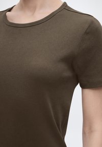 Κλείσιμο εικόνας ενός ατόμου που φοράει εφαρμοστό, κοντομάνικο, ριμπ ύφασμα σε σκούρο ελιά με στρογγυλή λαιμόκοψη.