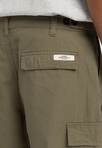 Olivgrön cargo shorts i lätt tyg, med sidoficka med lock, etikettpatch och synliga sömnadsdetaljer.